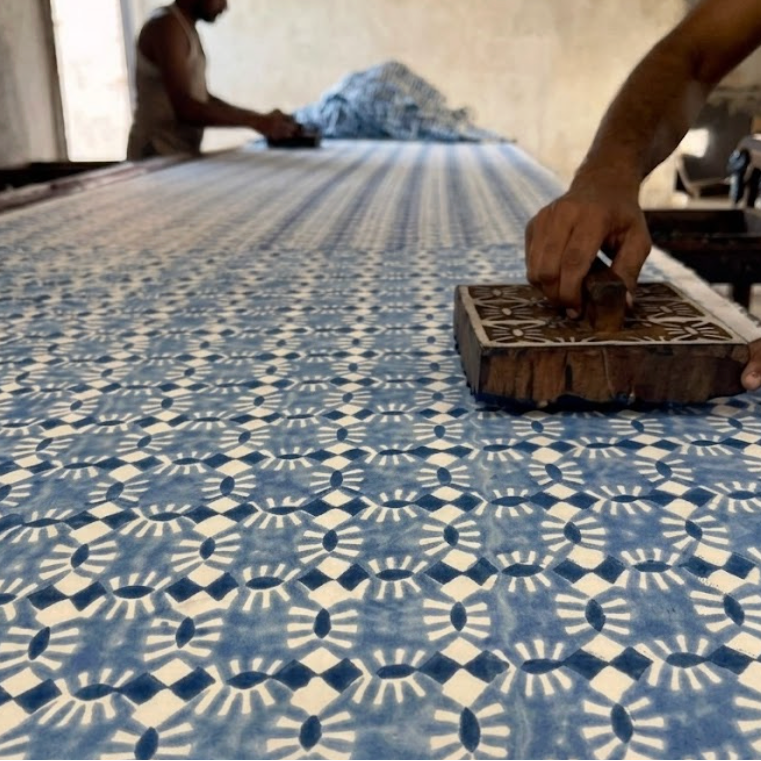 A close-up of a blue and white patterned fabric being printed or pressed with a wooden block in a workshop, with a person's hand in the foreground and another person visible in the background.