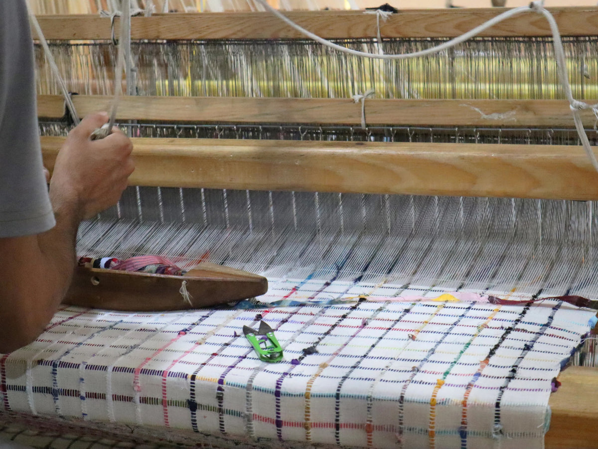 A close-up of a hand weaving a plaid textile on a wooden loom, with colorful threads and a white woven fabric visible.